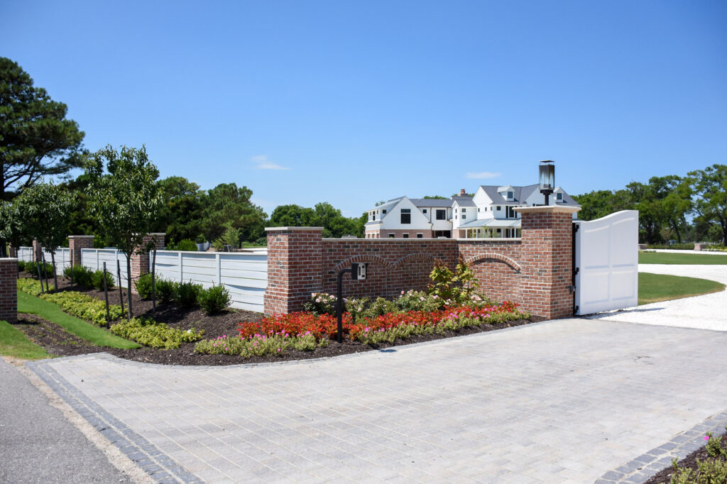 The entrance to a large estate with a big house in the background behind an automatic residential gate, a brick decorative wall, and a vinyl privacy fence.